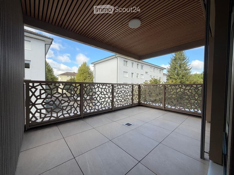 Balcony with tiled floor, decorative metal railing, wooden ceiling, and view of surrounding buildings and trees.