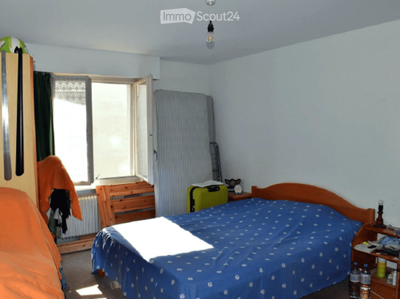 Bedroom with a blue bedspread, wooden nightstand, wardrobe, chair, suitcase, radiator, and a lamp on the nightstand.