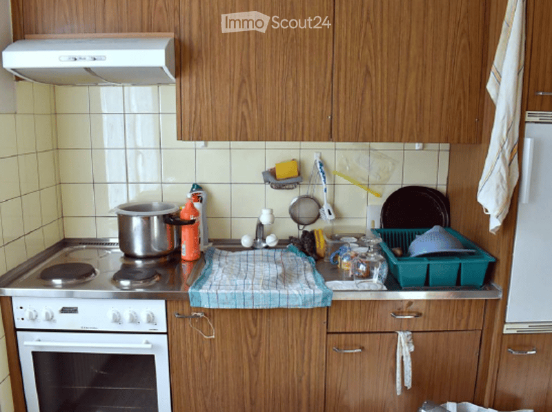 A small kitchen with wooden cabinets, a white stove, and a sink. Various kitchen items, including a pot, dish soap, and a dish rack, are visible on the counter.