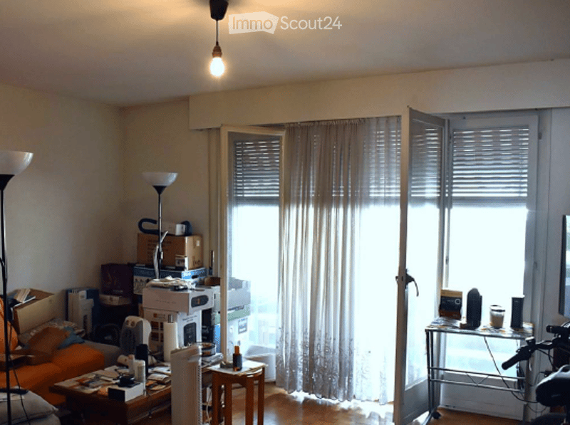 A furnished living room with a sofa, coffee table, floor lamp, small table with a chair, microwave, fridge, laptop, and various items scattered around. The room has large windows with curtains and wooden flooring.