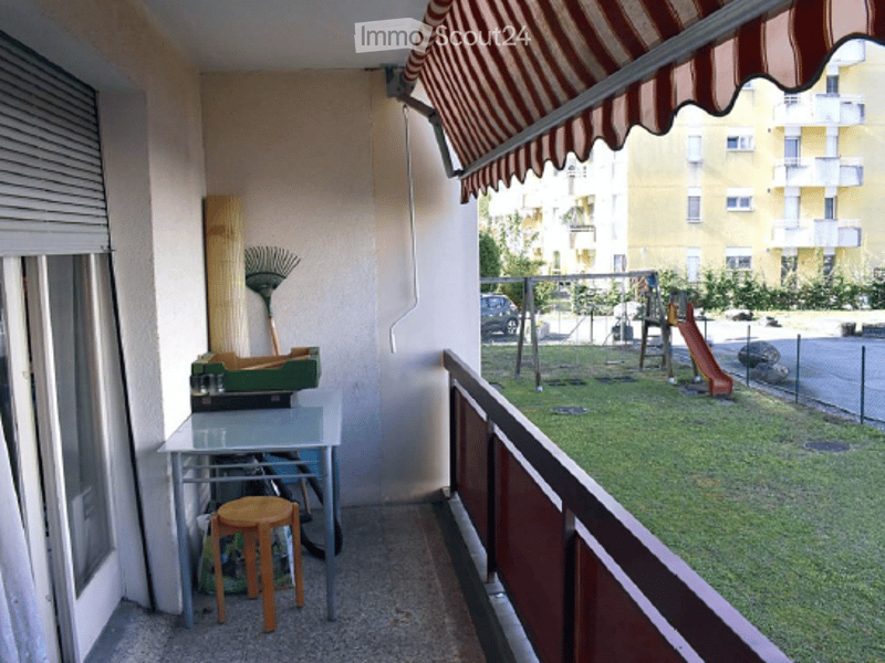 Balcony with a small table, stool, broom, and a view of a playground and residential buildings.