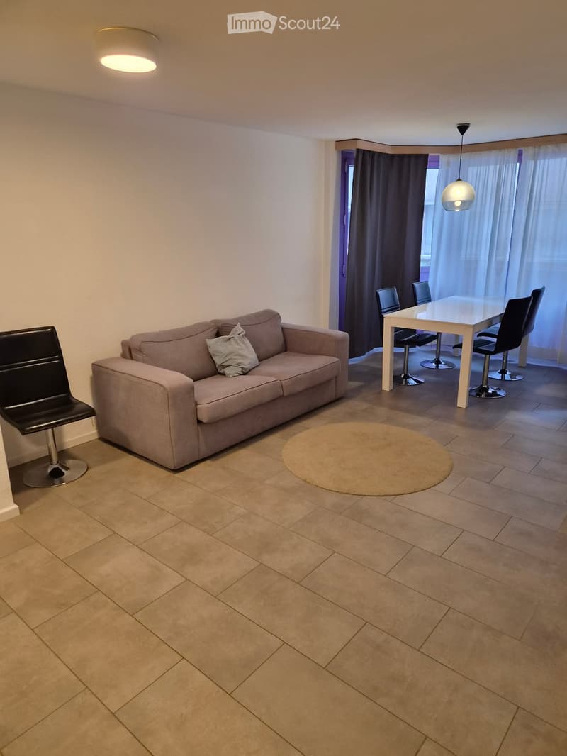 Living and dining area with a beige sofa, a round rug, a white dining table with four chairs, and a black bar stool near the sofa.
