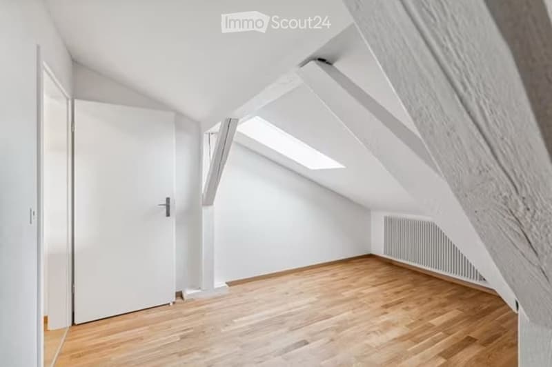 Bright attic room with wooden flooring, white walls, exposed wooden beams, and skylight windows. No furniture or equipment present.