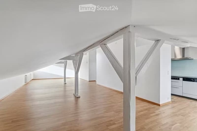 Open, loft-style room with wooden flooring, white walls, exposed wooden beams, and a small kitchen area with white cabinets and stainless steel appliances.