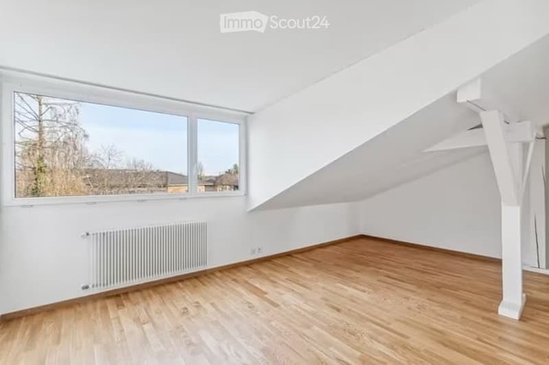 Completely empty room with wooden flooring, white walls, large window, radiator, and slanted ceiling.