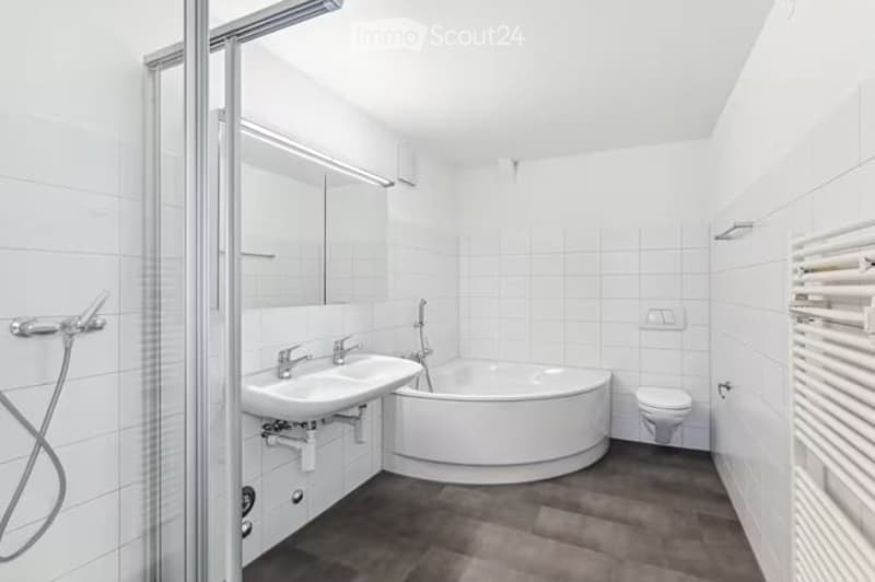 Modern bathroom with white tiles, a round bathtub, a sink, a toilet, a shower, and a mirror. The floor is dark grey, and there is a white radiator on the wall.