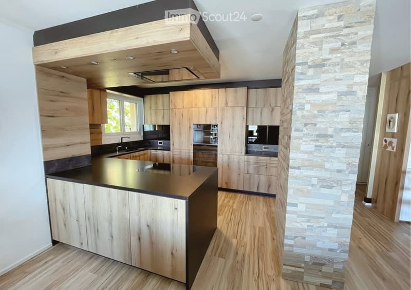 Modern kitchen with wooden cabinets, a central island, and a stone accent wall. Features include a black countertop, a sink, and a window providing natural light.
