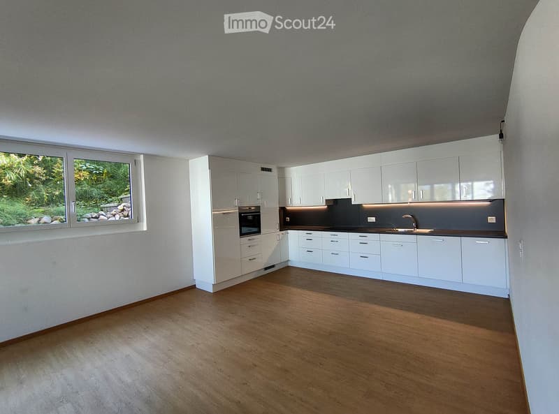 Modern kitchen with white cabinets, a sink, and an oven, featuring wooden flooring and large windows with a view of greenery.