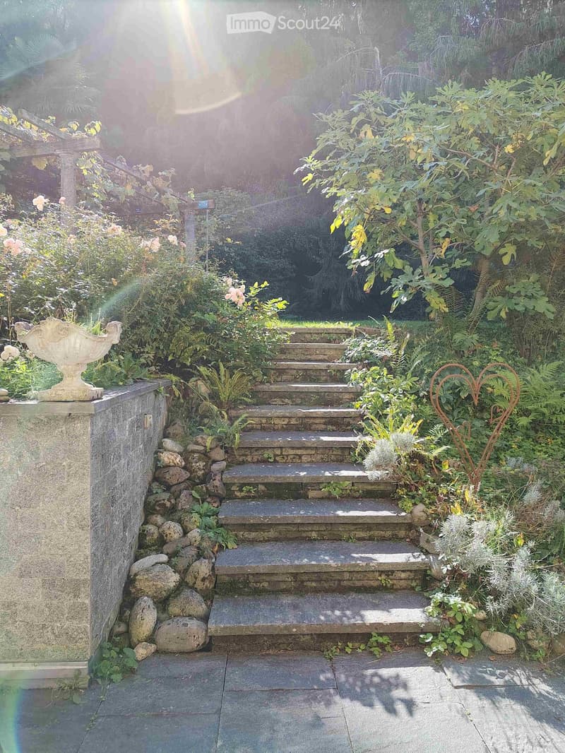 A lush garden with stone steps leading up, surrounded by greenery, flowers, and decorative elements like a stone urn and a heart-shaped metal sculpture.