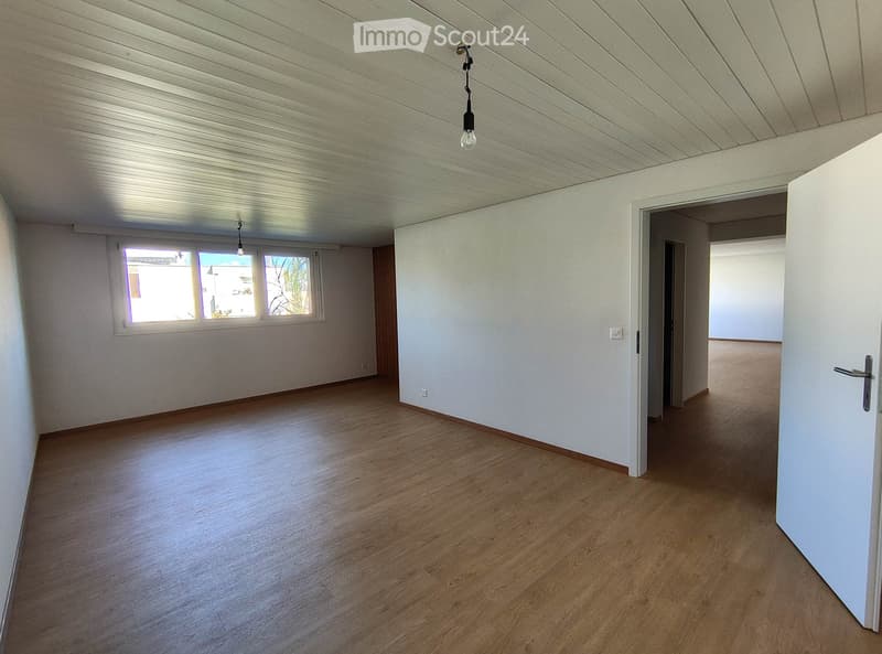Empty room with wooden flooring, white walls, and a slanted ceiling. Natural light enters through a window, and there is a doorway leading to another room.