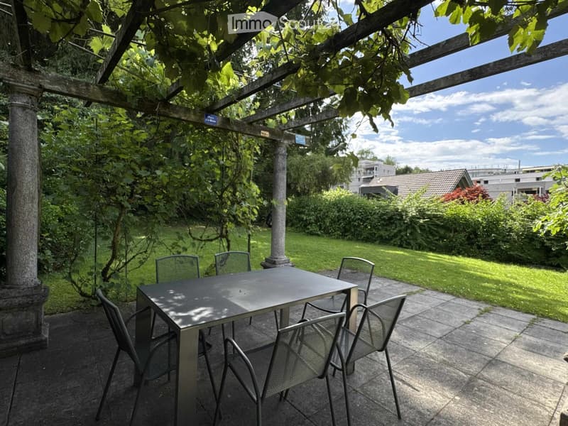 A covered outdoor patio with a table and chairs, surrounded by greenery and a well-maintained lawn.