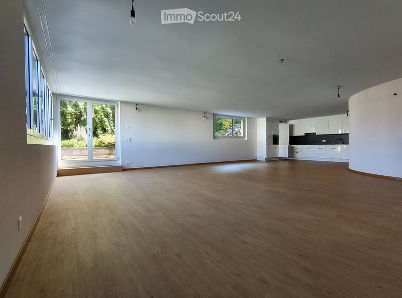 Empty room with wooden flooring, white walls, large windows, and a view of greenery outside. The room is unfurnished and has a modern, minimalist design.