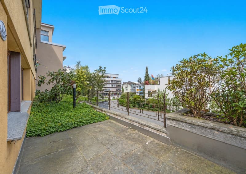 A paved outdoor area with low shrubs, small trees, and a metal railing leading to a pathway, surrounded by residential buildings.