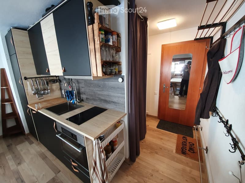 Modern kitchen with wooden countertops, black cabinets, sink, oven, and shelves holding jars and glasses. Wooden floor and a hallway with a coat rack and a welcome sign are visible.