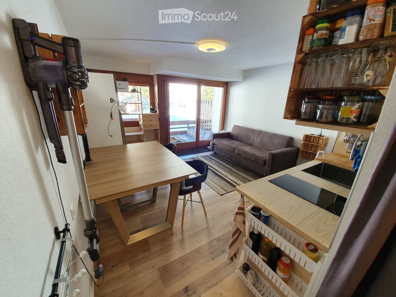 A modern, open-plan living and dining area with wooden flooring, a grey sofa, a wooden dining table with chairs, a kitchen counter with shelves, and a vacuum cleaner leaning against the wall.