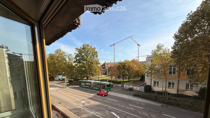 View from a balcony showing a street scene with a bus, a red car, trees, and construction cranes in the background.
