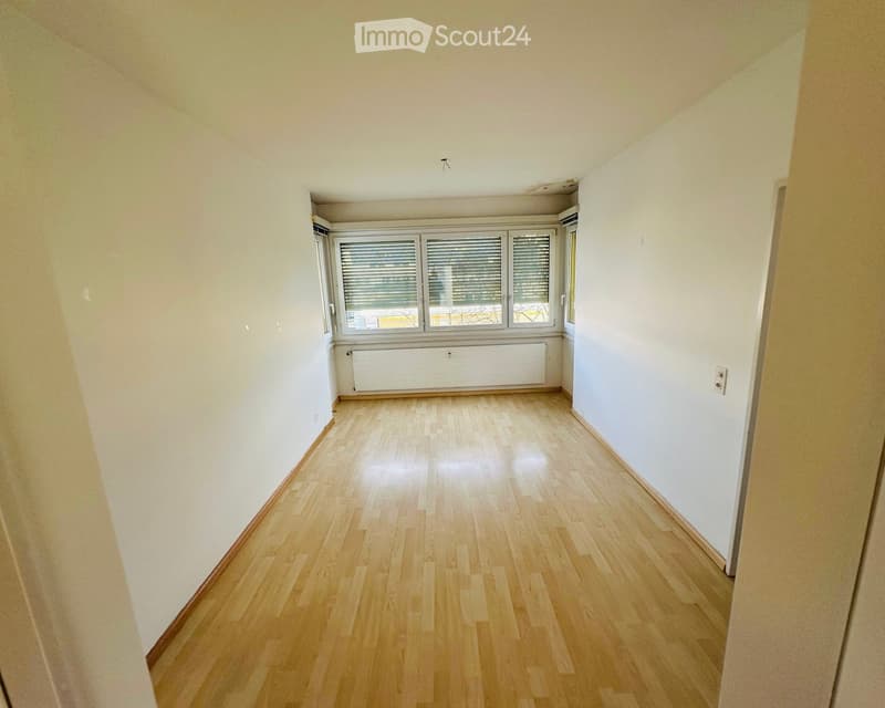 Empty hallway with wooden flooring, white walls, and large windows with blinds at the end.