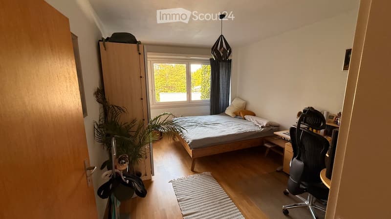 Cozy bedroom with wooden flooring, large window with dark curtains, bed with pillows and blanket, small table with chair, and potted plants.