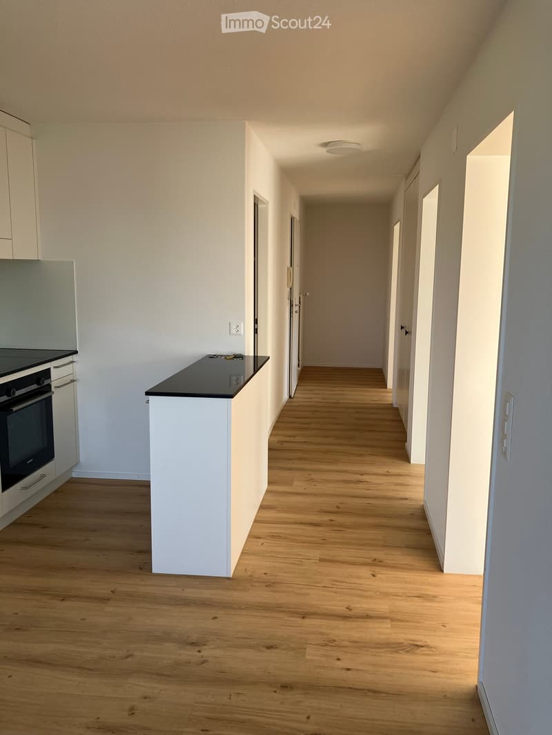 Empty hallway with wooden flooring, white walls, and a small kitchen area visible on the left side.