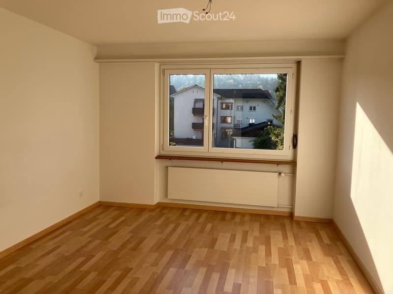 Empty room with wooden flooring, white walls, a large window with a view of neighboring houses, and a radiator below the window.
