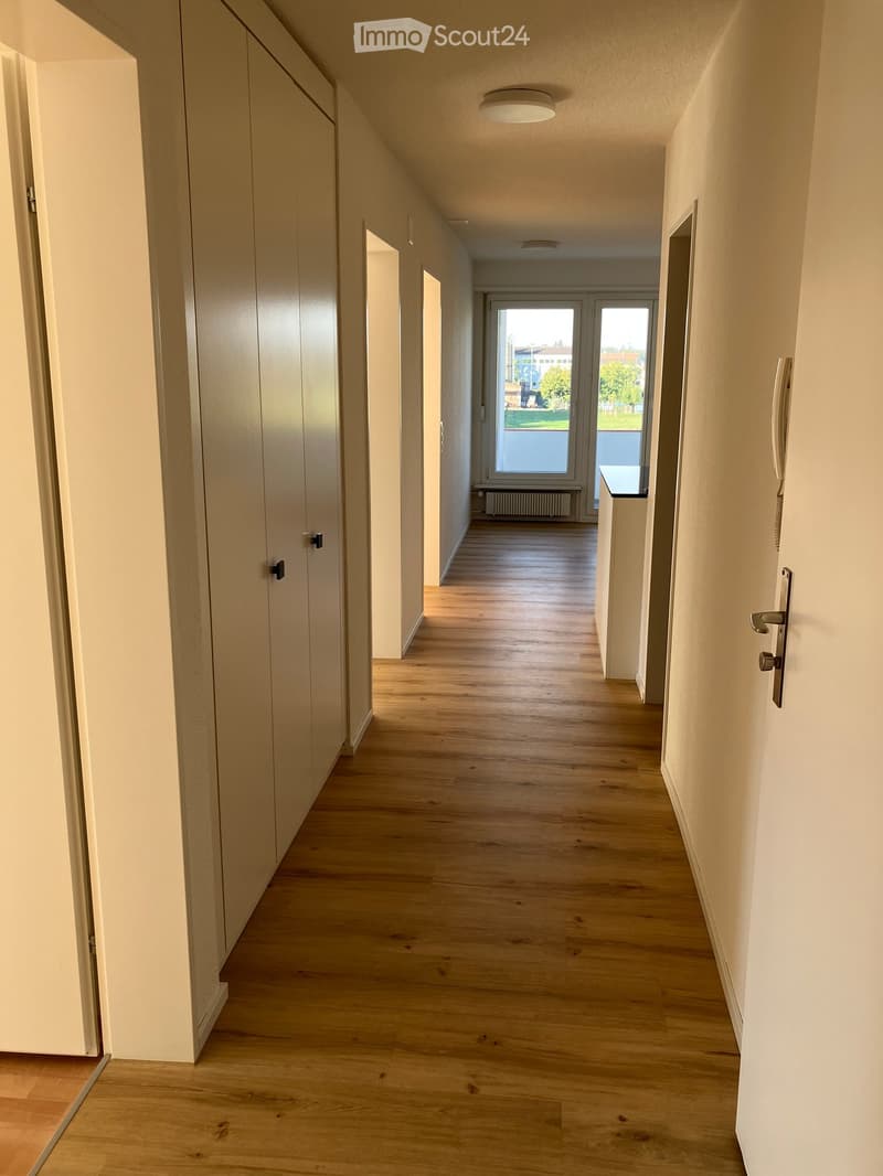 Bright hallway with wooden flooring, white walls, and several doors on both sides. Natural light streams in from a large window at the end of the corridor.