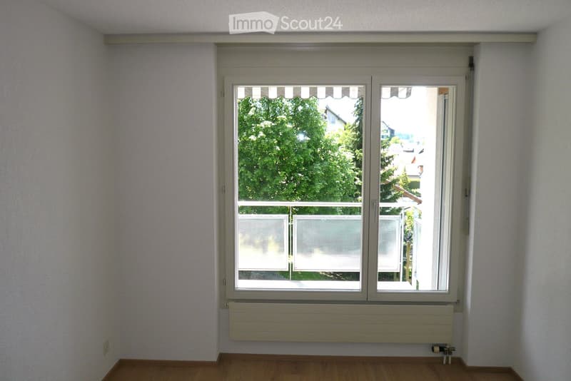 Empty room with wooden flooring, white walls, large window with blinds, radiator below the window, and a view of greenery and neighboring buildings.
