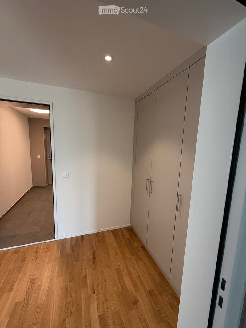 Empty hallway with light wooden flooring, white walls, built-in closet with mirrored doors, and recessed ceiling lighting.