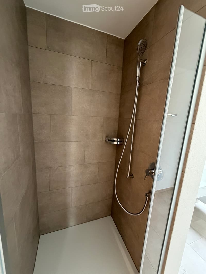 Modern shower area with beige tiled walls and floor, featuring a handheld showerhead and a fixed showerhead, enclosed by a glass shower door.