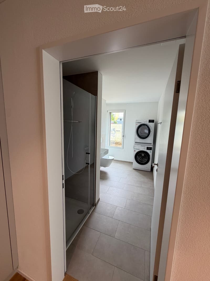 Modern bathroom with a glass-enclosed shower, toilet, and stacked washing machine/dryer unit. Light-colored tiles and neutral walls.