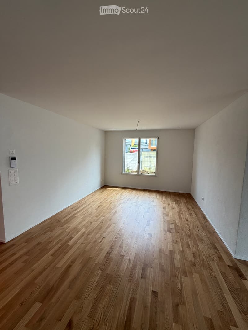 Empty room with wooden flooring, white walls, and a large window letting in natural light.