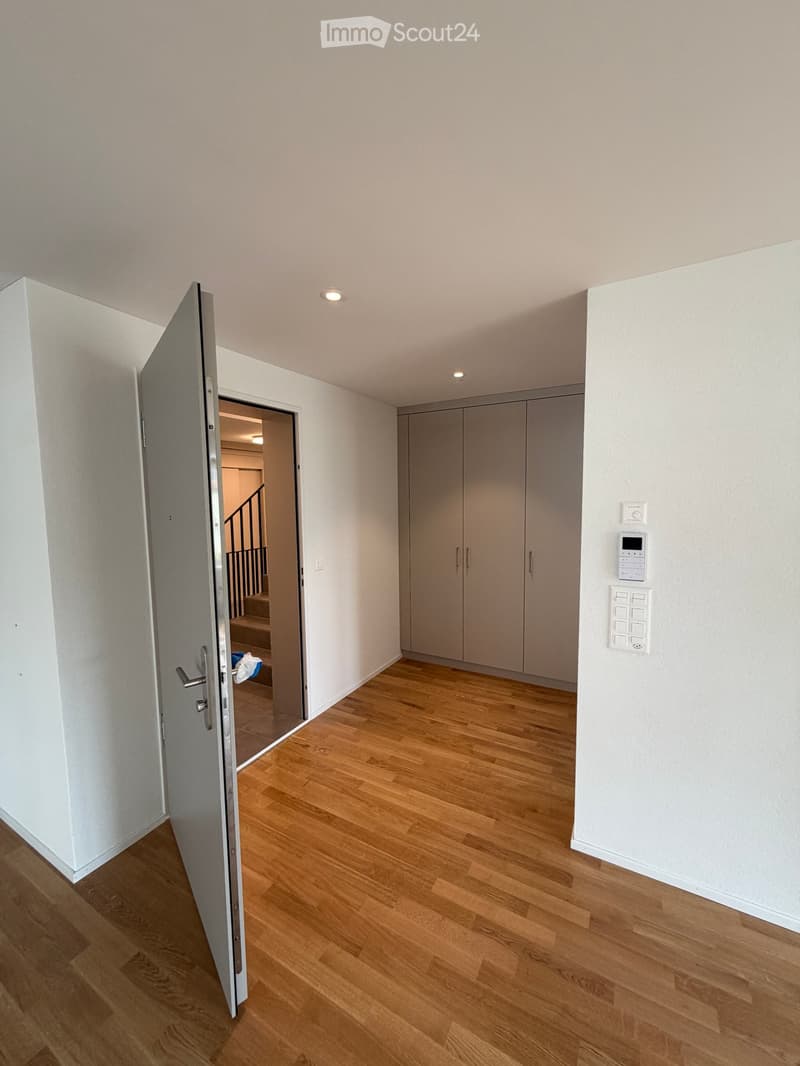 Empty hallway with wooden flooring, white walls, and a partially open door leading to another room. There are built-in cabinets and a control panel on the wall.
