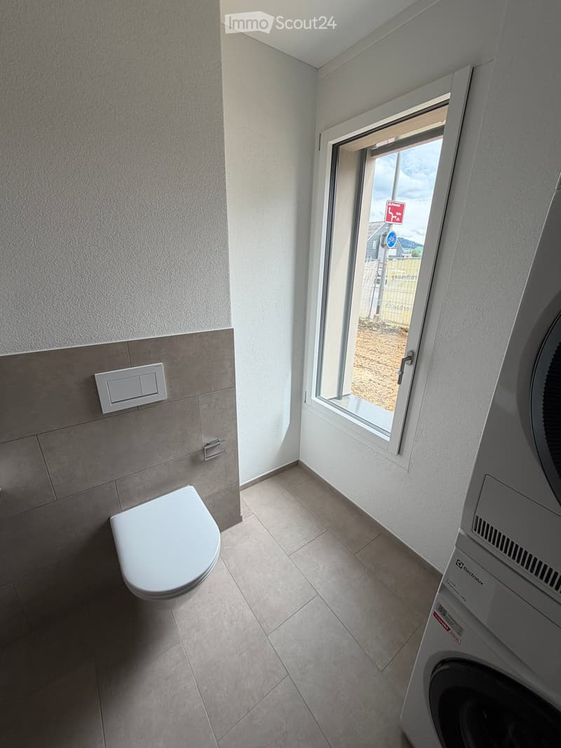 Modern bathroom with a toilet, washing machine, tiled walls, and a window offering a view of the outdoor area.