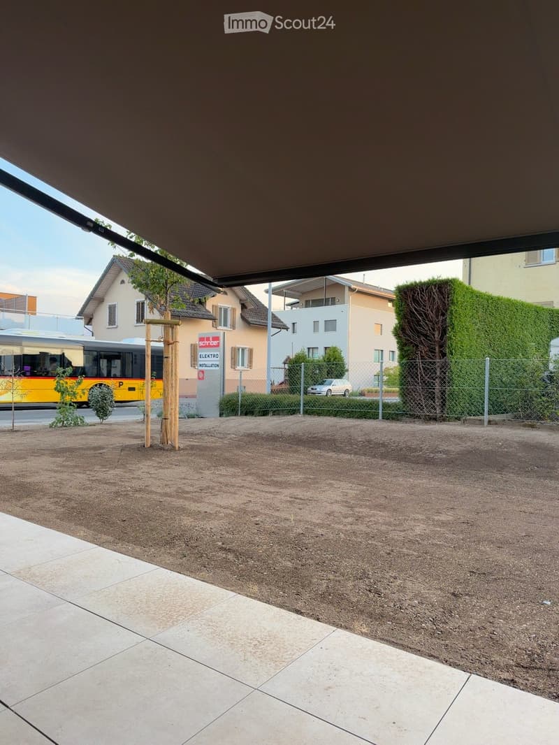 Outdoor area with gravel ground, tiled walkway, small wooden structure, fence, and surrounding buildings.