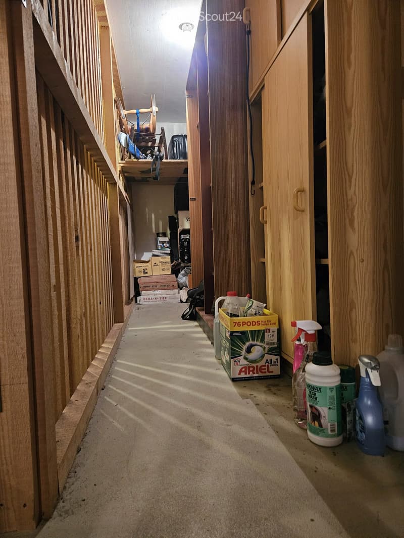 A narrow, unfinished hallway with wooden paneling on one side and exposed concrete flooring. Cleaning supplies and boxes are placed along the walls.
