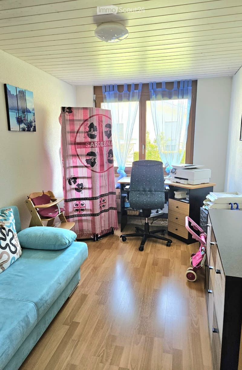 A multi-functional room with a sofa, office chair, desk, printer, baby chair, and baby stroller. The room features wooden flooring, large windows with curtains, and a picture frame on the wall.