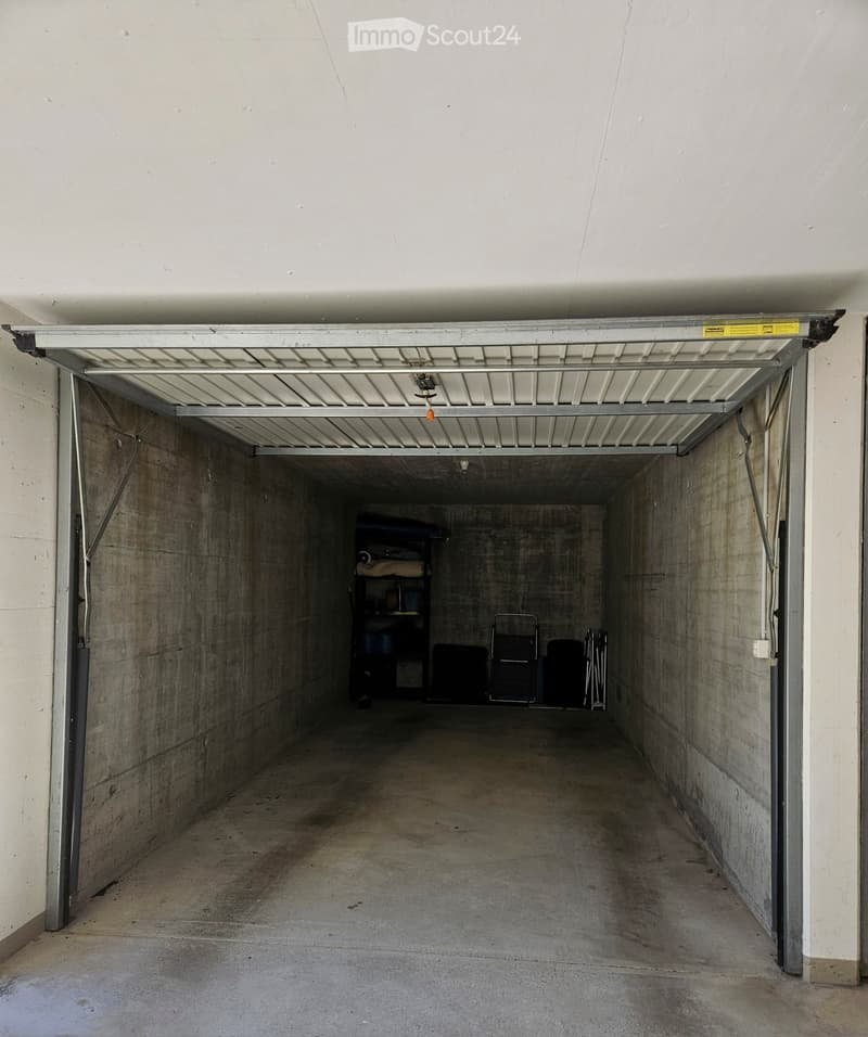 Underground garage with concrete walls and floor, open garage door, and some stored items including a shelf and chairs.