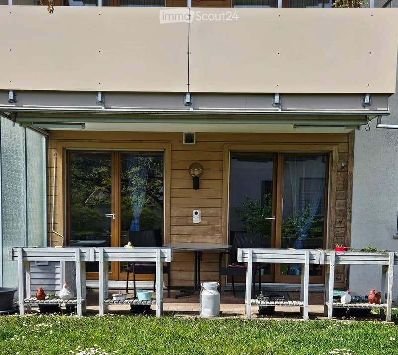 Balcony area with a table, chairs, benches, potted plants, and decorative items, featuring a covered terrace and glass doors leading inside.