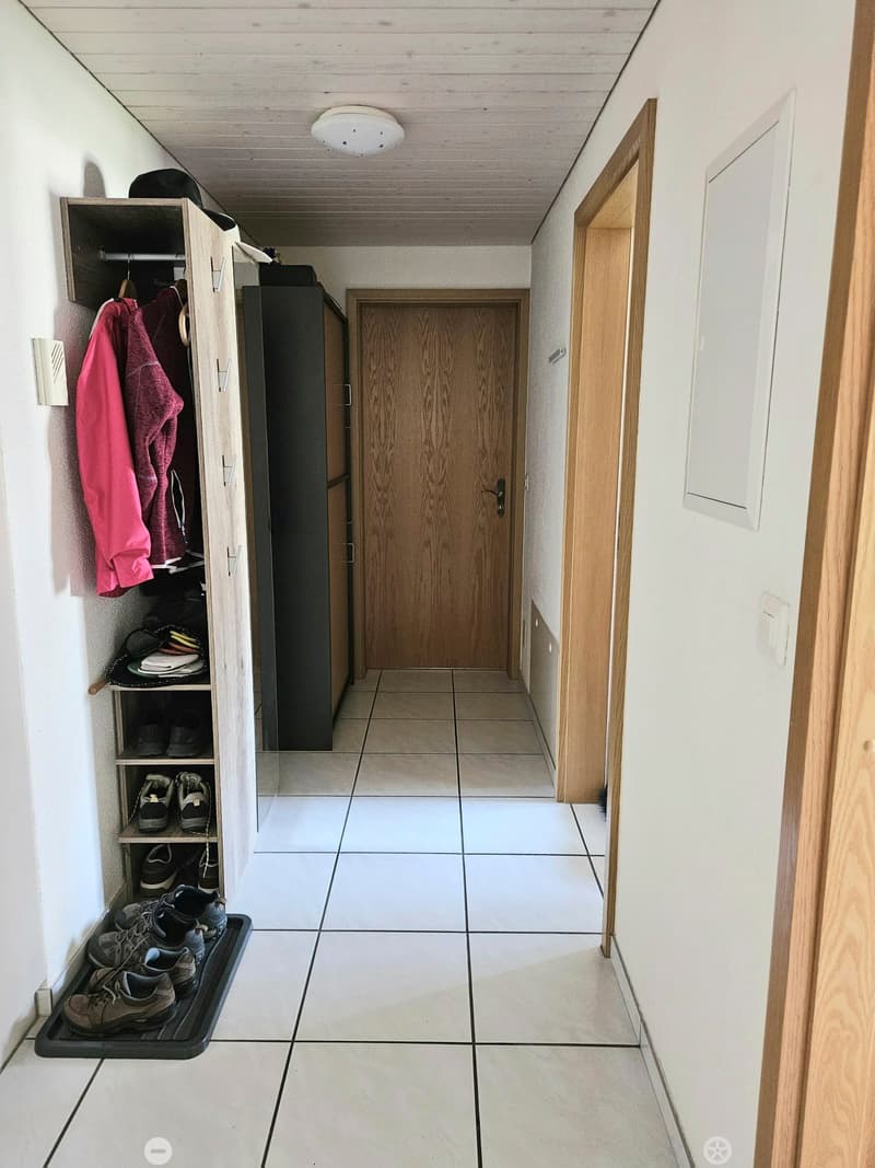 A narrow hallway with tiled flooring, white walls, and wooden doors. A shoe rack with multiple pairs of shoes and a coat rack with hanging clothes are visible on the left side.