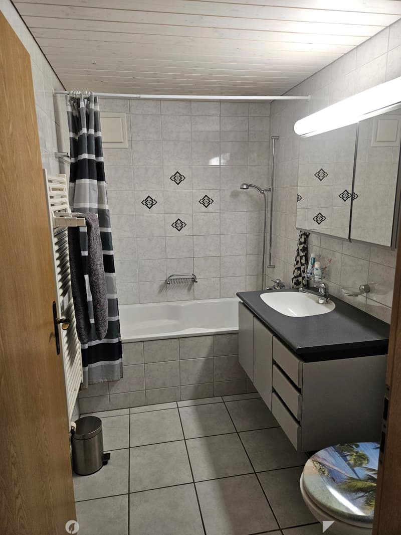 Bathroom with a bathtub, sink, toilet, mirror, and shower curtain. Features tiled walls and floor, a wooden door, and a towel hanging on a rack.