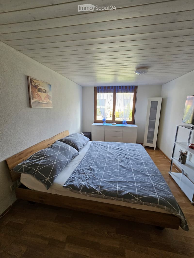 Cozy bedroom with wooden bed, nightstand, dresser, and cabinet. Features a large window with blue curtains and a picture frame on the wall.