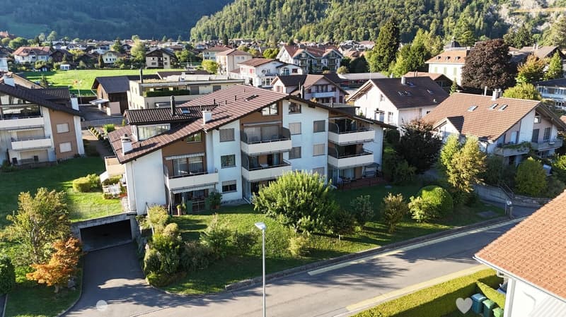 Aerial view of a multi-story residential building with a brown tiled roof, surrounded by greenery and neighboring houses in a suburban area.