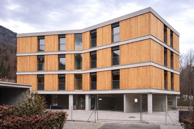 3 story wooden-paneled building, windows with balcony, fenced off entrance, ground floor with concrete pillars