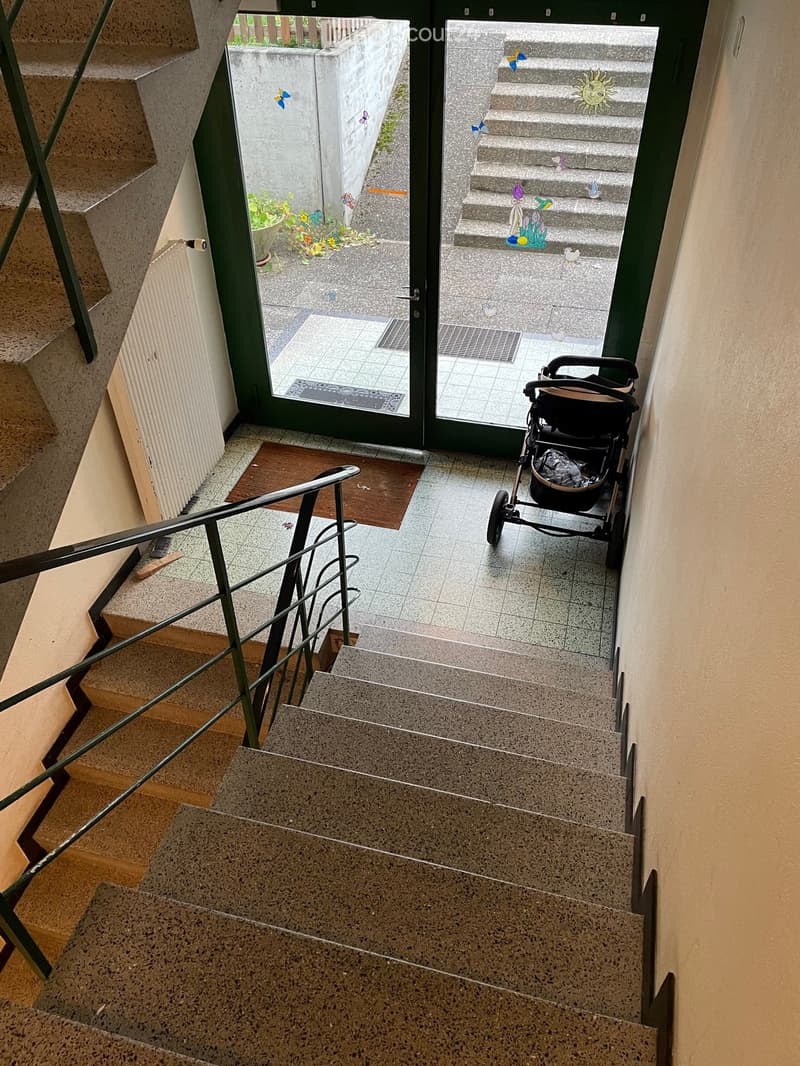 Staircase leading to an outdoor area with a stroller visible near the entrance. The hallway has tiled flooring and a metal railing.