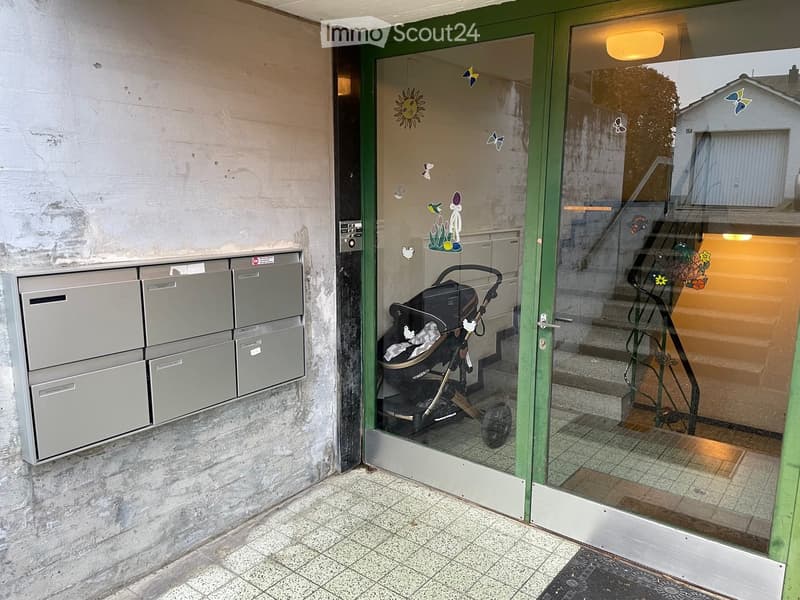 Entrance area with mailboxes on the left and a stroller visible through the glass door, leading to a staircase inside the building.