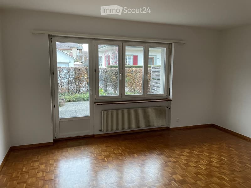 Empty room with wooden flooring, large windows, radiator, and a sliding door leading to an outdoor area.