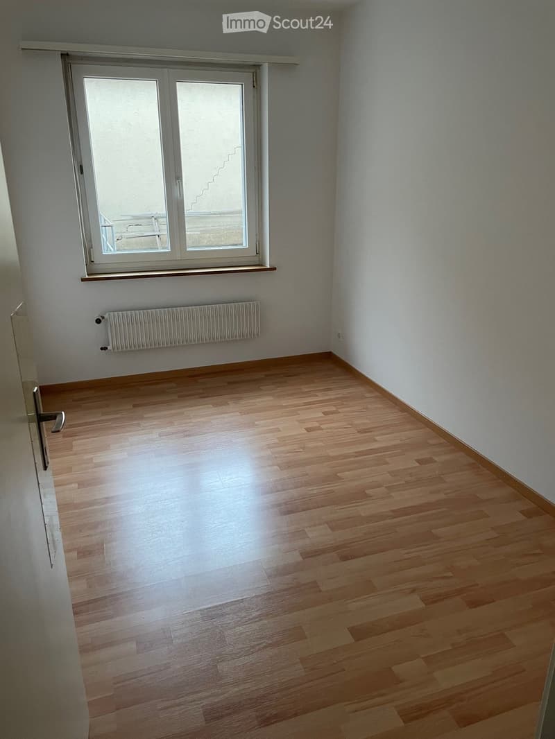 Empty room with wooden flooring, white walls, a window with a radiator below, and a door visible on the left side.