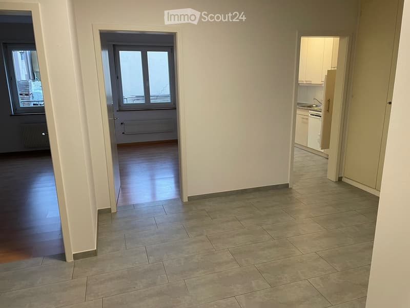 Empty hallway with tiled floor and wooden flooring in adjacent rooms, featuring white walls and large windows.