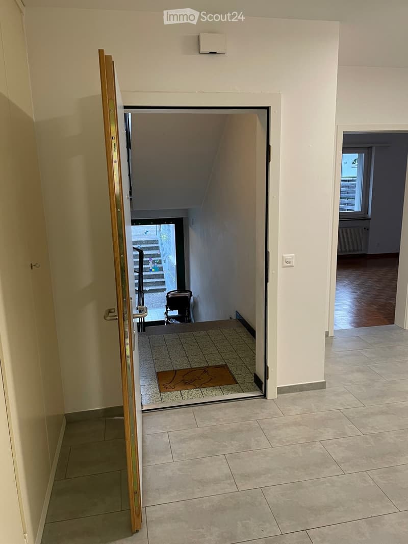 Hallway with tiled floor, open door leading to a staircase area with a stroller visible, and another room with wooden flooring visible through an open doorway.