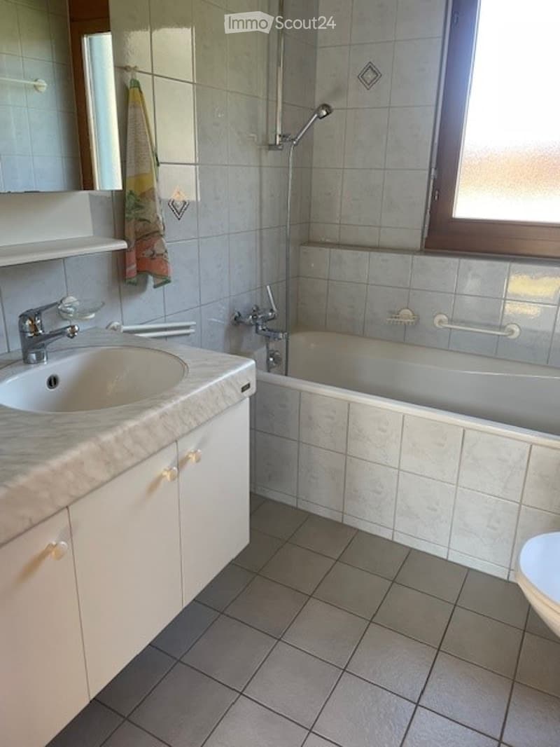 Bathroom with a white tiled wall, a marble countertop sink, a bathtub, a mirror, and a handrail for safety.