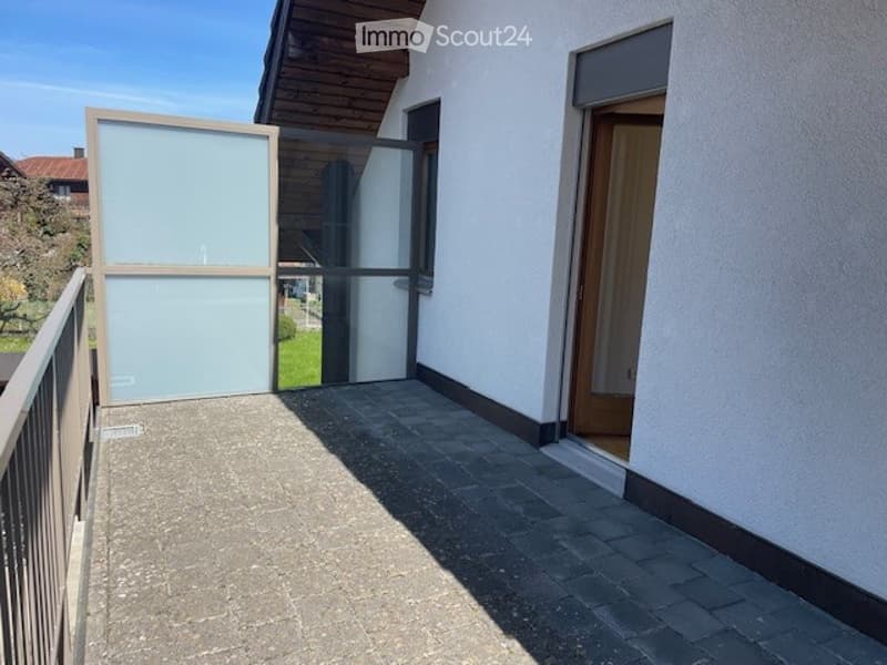 Balcony with a modern glass door, metal railing, and tiled floor, leading to a wooden door of the house.